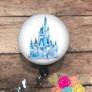 May include: A round, white retractable badge reel featuring a watercolor image of a blue castle. The castle has multiple spires and is set against a background of blue and purple watercolor splashes. The reel has a black clip and a clear plastic strap.