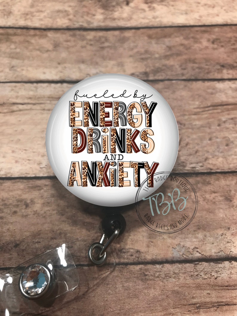 May include: A white round badge reel with a black retractable cord and a clear plastic badge holder. The badge reel features a leopard print design with the text "fueled by energy drinks and anxiety".
