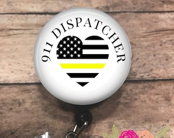 Op de afbeelding: Een witte badge reel met de tekst "911 DISPATCHER" rond een hartvormig ontwerp. Het hart heeft een zwart-wit Amerikaanse vlag met een gele streep. De reel heeft een zwarte clip en een doorzichtige plastic band.