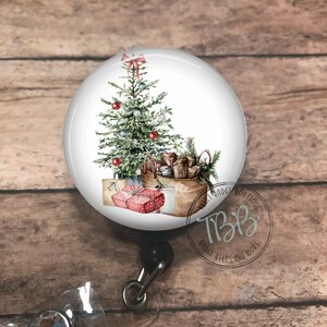 May include: A round badge reel with a Christmas tree design. The image features a decorated evergreen tree with red ornaments, gifts, and a basket of presents. The reel has a clear plastic strap and a decorative gem.