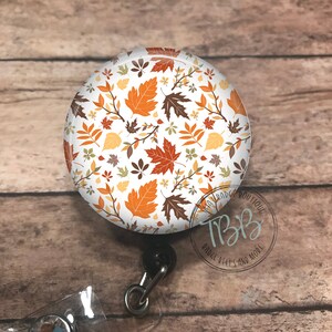 May include: A round, white badge reel with a fall leaf pattern. The leaves are in shades of orange, yellow, brown, and red. The badge reel has a clear plastic badge holder and a silver swivel clip.