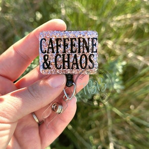 May include: A pink glitter badge reel with a black clip and a black and white square charm that says "CAFFEINE & CHAOS".