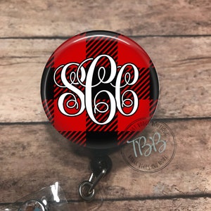 May include: A red and black buffalo plaid patterned badge reel with a white monogram. The badge reel is attached to a clear plastic badge holder with a crystal.