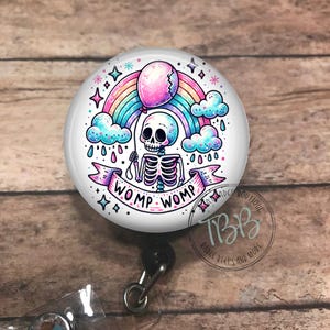 May include: A white badge reel with a colorful cartoon skeleton holding a broken pink balloon. The skeleton is standing in front of a rainbow with clouds and raindrops. The text "Womp Womp" is written on a banner below the skeleton.
