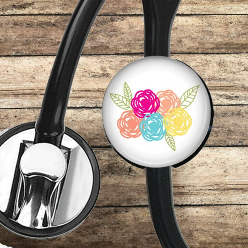 May include: A black stethoscope with a white round charm featuring a colorful floral design. The charm has five pink, orange, blue, and yellow flowers with green leaves.