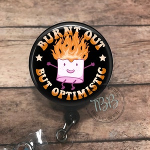 May include: A black button badge with a white marshmallow character with orange flames for hair. The text on the badge reads "Burnt Out But Optimistic".