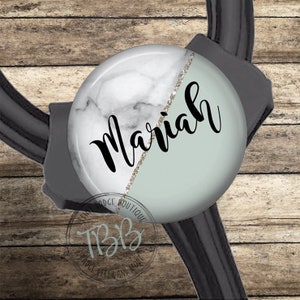 May include: A white and light blue marble stethoscope charm with gold glitter accents and the name "Mariah" written in black cursive.