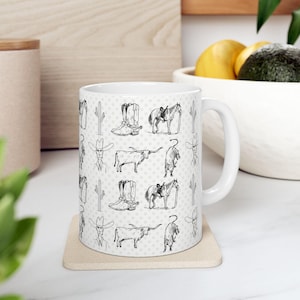 Może przedstawiać: Biały ceramiczny kubek z powtarzającym się wzorem ilustracji o tematyce kowbojskiej w czerni i bieli. Projekt obejmuje kowbojskie buty, konie, kaktusy, bydło długorogie i kowbojów. Kubek stoi na beżowej podstawce.