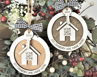 May include: Two white wooden ornaments with a brown woodgrain design. Each ornament features a house silhouette with the text "Home Sweet Home" and a key with the year "2023" on it. The bottom of each ornament has the text "1225 Cranberry Lane, Maine 04090".