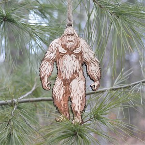 May include: Wooden Bigfoot ornament with a jute string for hanging. The ornament is a light brown color and features detailed etched lines to depict the creature's fur and facial features. The ornament is hanging from a pine tree branch.