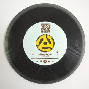 Puede incluir: Un disco de vinilo negro con una etiqueta blanca que presenta un adaptador de 45 RPM amarillo y naranja, un código QR y el texto "A Night Like This" de Caro Emerald. El disco tiene un botón de reproducción, un botón de pausa y un botón de retroceso.
