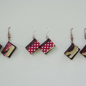 Può includere: Tre paia di orecchini a forma di libro. Il primo paio è rosso con pois bianchi, il secondo paio è floreale rosa e bianco, e il terzo paio è marrone con cerchi neri e bianchi.