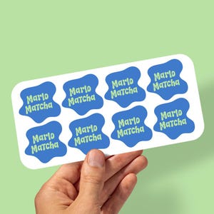 May include: A rectangular white sticker sheet featuring eight individual blue stickers with irregular, wavy edges. Each blue sticker displays the text Marlo Matcha printed in a light green, rounded, bubbly font across two lines.
