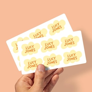 May include: Two white rectangular sheets of stickers are held in a hand. Each sticker features the name "LUCY JONES" in orange text within a yellow cloud shape. The background is a solid peach colour.