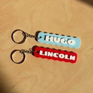 May include: Two personalised keyrings. One is light blue with the name "HUGO" in white, and the other is red with "LINCOLN" in white. Both have a wavy edge design and a silver keyring.