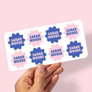 May include: A sheet of eight stickers with the name "SARAH WOODS" printed in a playful font. The stickers alternate between blue and pink starburst shapes on a white background. The background is a soft pink.