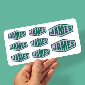 May include: A sheet of stickers featuring the name "JAMES" in a green and lavender geometric design. The stickers are held by a hand against a teal background. The name is in a bold, sans-serif font.