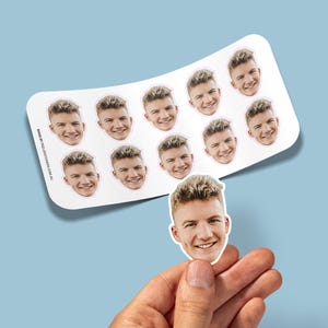 May include: A sheet of stickers featuring a smiling face with light brown hair. The stickers are white with a pink border and are held by a hand against a light blue background. The text "MADE BY PEELSTICKERS.COM.AU" is printed on the sheet.