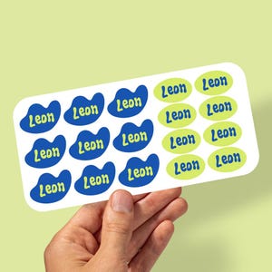May include: A sheet of stickers featuring the name "Leon". The stickers are in two shapes: blue blobs and green ovals. The name is printed in a contrasting colour on each sticker, suitable for labelling items.
