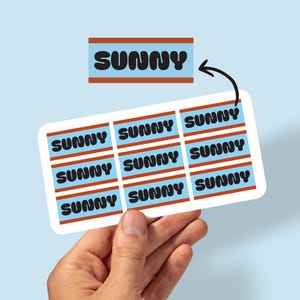 May include: A sheet of stickers with the word "SUNNY" in bold black letters on a light blue background, framed by a brown border. The stickers are held by a hand against a light blue backdrop.