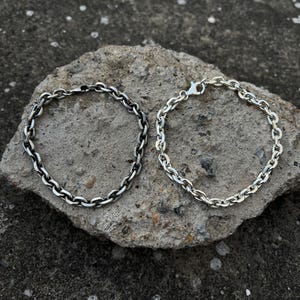 May include: Two silver chain bracelets with a linked design. One bracelet has a darker, oxidized finish, while the other is bright silver. The bracelets are displayed on a rough, gray stone surface.