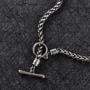 May include: Close-up of a silver-toned chain necklace with a toggle clasp. The chain has a braided design, and the clasp features a circular ring and a cylindrical bar. The necklace is displayed against a dark, textured background.