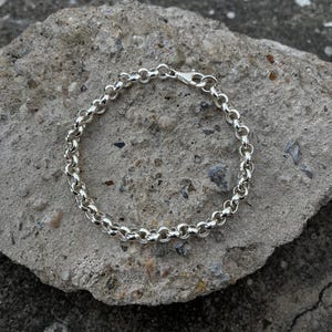 Puede incluir: Una pulsera de cadena de plata con cierre de mosquetón. La pulsera está compuesta por pequeños eslabones redondos e interconectados. La joya se exhibe sobre una superficie de piedra gris rugosa.