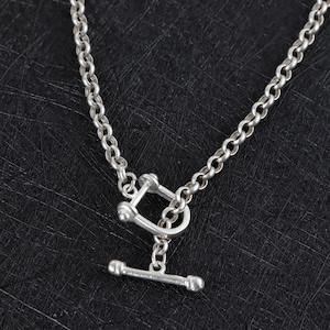 May include: A silver-toned necklace with a toggle clasp. The chain is made of small, rounded links. The clasp features a rectangular loop and a horizontal bar with rounded ends. The necklace is displayed against a dark background.