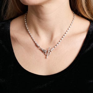 Puede incluir: Un collar plateado con pequeñas cuentas ovaladas. El cierre presenta dos manos que sostienen una barra. El collar se muestra sobre un fondo negro. Una joya única y elegante.