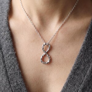 Può includere: Un ciondolo a forma di simbolo dell'infinito color argento con una superficie martellata e testurizzata, sospeso a una catenina a maglia rolo abbinata. Il ciondolo presenta un design a ciclo continuo con un aspetto metallico riflettente.