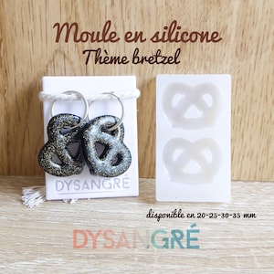 Puede incluir: Dos llaveros con forma de pretzel negros y dorados con purpurina y anillos plateados, expuestos sobre una tarjeta blanca. También se muestra un molde de silicona blanco con dos formas de pretzel. El texto "Moule en silicone Thème bretzel" está en la parte superior.