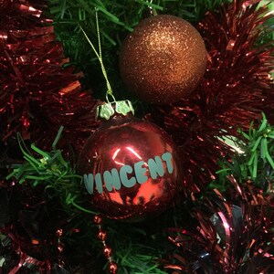 May include: A red Christmas ornament with the name "Vincent" written on it. The ornament is hanging on a Christmas tree with red and green tinsel.