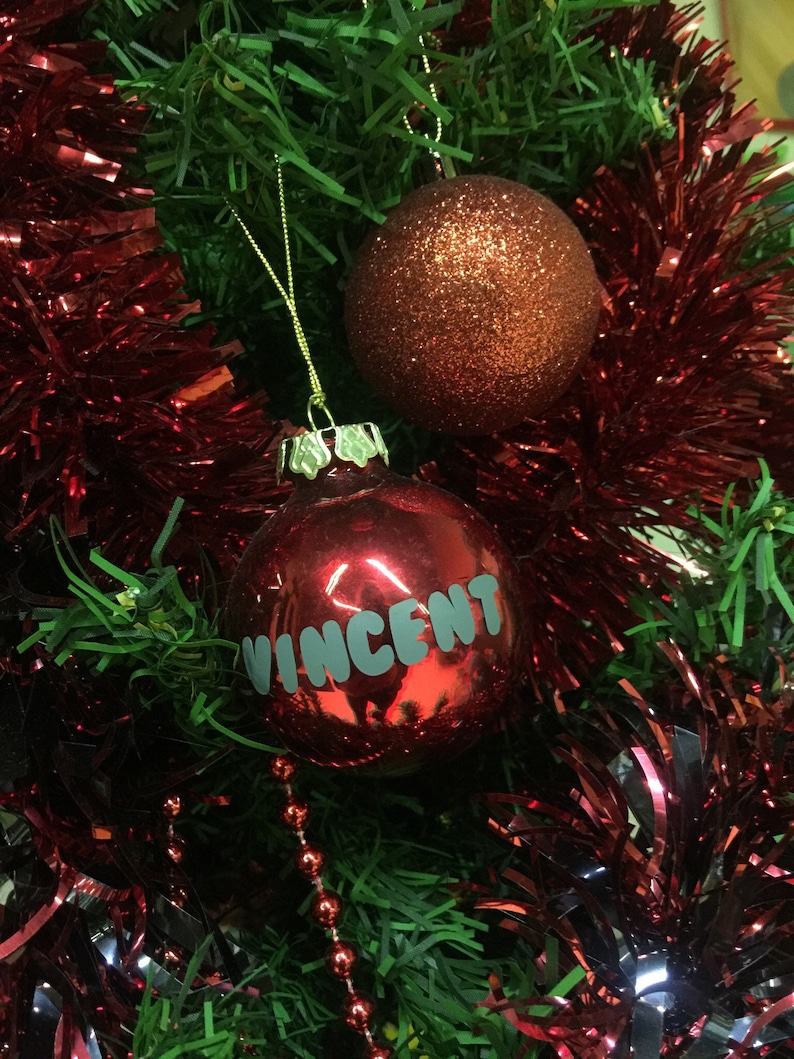 May include: A red Christmas ornament with the name "Vincent" written on it. The ornament is hanging on a Christmas tree with red and green tinsel.