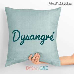 以下が含まれることがあります： 薄い青色のクッションで、前面に濃い青色の糸で「Dysangré」と刺繍されています。