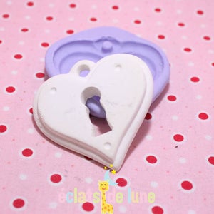 Puede incluir: Un colgante blanco en forma de corazón con un recorte en forma de ojo de cerradura. El colgante está hecho de un material liso y mate y tiene un pequeño bucle en la parte superior para colgar. El colgante está sentado sobre un fondo de lunares rosa y blanco.