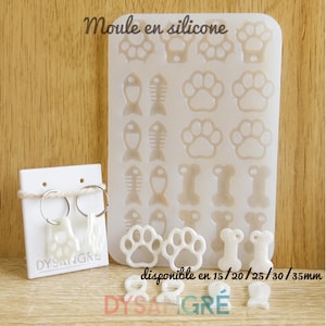 Può includere: Uno stampo in silicone bianco con impronte di zampe, lische di pesce e forme di ossa di cane. Un paio di orecchini a forma di impronta di zampa sono esposti su una carta bianca. Il testo "Moule en silicone" è in alto.