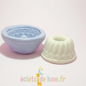 Puede incluir: Un molde de silicona de color morado claro para hacer un pequeño pastel o postre. El molde se muestra junto a un pastel blanco que se ha hecho utilizando el molde.