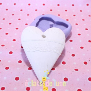 Puede incluir: Un adorno blanco en forma de corazón con un diseño de encaje y un agujero en la parte superior. El adorno está sobre una tela de lunares rosa y blanca.