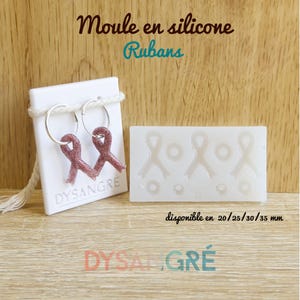 Pode incluir: Brincos de argola com pingentes de fita e um molde de silicone para fazer formas de fita. Os brincos são de cor bordô brilhante. O molde é branco e tem três formas de fita e três círculos. O texto "Moule en silicone Rubans" está no topo.