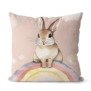 May include: A decorative square pillow featuring a watercolor illustration of a brown bunny sitting on a rainbow. The background is a soft pink with white cloud-like accents. The pillow is a home decor item.