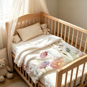 Puede incluir: Una cuna de madera que contiene una almohada blanca, un oso de peluche y una manta blanca suave con un estampado de acuarela de flores silvestres rosas, moradas y verdes. La cuna está cubierta con un dosel blanco, creando un espacio luminoso.