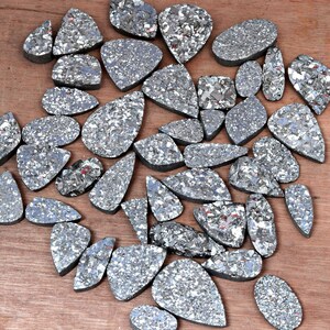 May include: A collection of silver and gray gemstone cabochons, each with a unique, irregular shape and a sparkly, iridescent finish. The stones are arranged in a random pattern on a wooden surface.