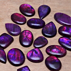 May include: A collection of 15 purple and pink labradorite gemstone cabochons. The cabochons are various shapes and sizes, with some having a pear shape and others being oval.