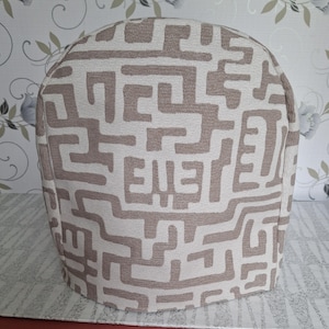 Op de afbeelding: Een afgeronde theemuts van stof met een getextureerde gebroken witte basis en een herhalend taupekleurig, doolhofachtig geometrisch patroon. De hoes heeft een gestructureerd, koepelvormig silhouet ontworpen voor een theepot van geweven textiel.