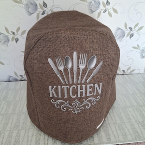 Può includere: Una copertura marrone per elettrodomestici da cucina con un design di posate argentate e la parola "KITCHEN" in lettere argentate. La copertura è realizzata in tessuto strutturato e presenta un motivo a volute decorative.