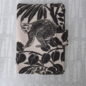 Peut inclure: Étui en tissu beige et noir avec un motif de lémurien entouré de feuilles tropicales. Le lémurien est représenté dans un style artistique détaillé. L'étui est doté d'une fermeture à rabat, probablement pour une tablette ou un ordinateur portable.