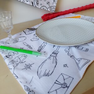 Peut inclure: Un set de table blanc avec une illustration en noir et blanc d'un balai, d'une plume, d'une fiole de potion et d'une enveloppe. Un marqueur vert est posé sur le set de table.