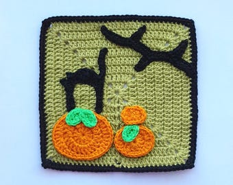 May include: A crocheted square featuring Halloween-themed elements. The design includes a black cat, two orange pumpkins with green leaves, and a black tree branch against a green background. The square is outlined in black.
