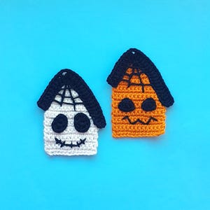 Könnte beinhalten: Zwei gehäkelte Halloween-Hausornamente. Eines ist weiß mit schwarzen Augen, einem Mund und einem schwarzen Dach mit Spinnennetz-Design. Das andere ist orange mit schwarzen Augen, einem Mund und einem schwarzen Dach mit Spinnennetz-Design. Der Hintergrund ist einfarbig hellblau.