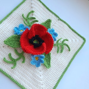 Pode incluir: Um quadrado branco de crochê com uma borda verde apresenta uma flor de papoula vermelha com um centro preto rodeado de flores azuis e amarelas de miosotis e folhas verdes.